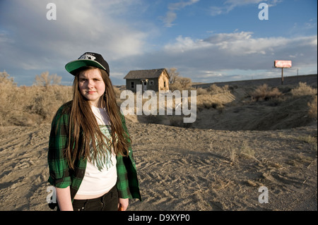 Teenage girl with long brown hair wearing an OBEY cap and green check shirt standing in scrub land with shack & sign behind her. Stock Photo