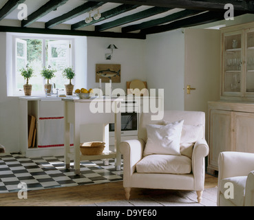 Black+white tiled floor in white country cottage kitchen open to a sitting area  with white armchair and lime washed dresser Stock Photo