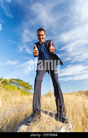 cheerful young businessman on top of rock giving thumbs up outdoors Stock Photo