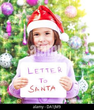 Closeup on cute little girl holding in hands letter to Santa Claus, wearing red festive hat, beautiful decorated Christmas tree Stock Photo