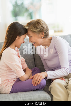 Granddaughter (8-9) and grandmother sitting on sofa and touching with heads Stock Photo