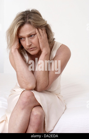 Thoughtful mature woman in nightdress sitting on bed Stock Photo