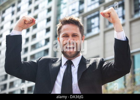 A young successful man, male executive businessman arms raised celebrating cheering shouting in front of a high rise office Stock Photo