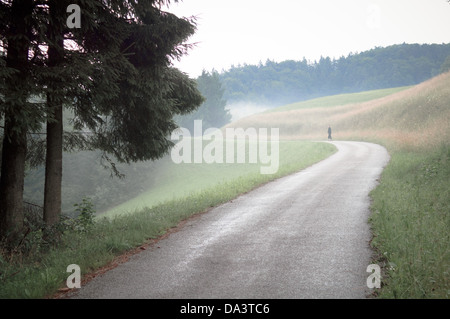 Woman walking alone on windy road uphill towards misty forest Stock Photo