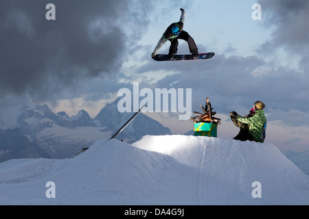 ZERMATT, SWITZERLAND. A freestyle snowboarder is executing a tailgrab trick and jumps over an oil barrel with a fire on it. Stock Photo