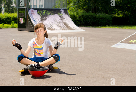 Pretty young teenage girl in rollerblades sitting meditating on the tarmac at a skate park sitting n the lotus position with her eyes closed. Stock Photo