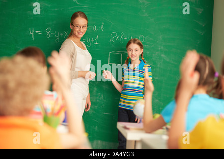 Portrait of smart teacher and schoolgirl standing by blackboard and looking at schoolkids in classroom Stock Photo