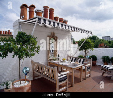 Black+white striped awning above wooden table and chairs set for lunch on roof terrace with bay trees in terracotta pots Stock Photo