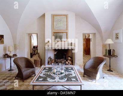 Pair of wicker chairs either side of fireplace in French country living room with tiled coffee table and vaulted ceiling Stock Photo