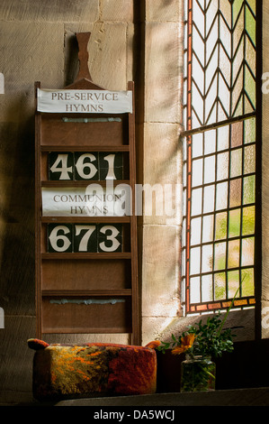 Close-up of stained glass church window with hymn board, kneeler & small vase of flowers on stone ledge - St. Mary's Church, Kettlewell, Yorkshire, UK Stock Photo