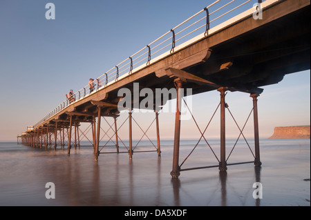 Blue summer evening sky & view of people standing & walking in sun, on historic scenic seaside pier over calm sea - Saltburn-by-the-Sea, England, UK. Stock Photo