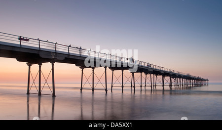 Under colourful summer sunset sky, view from sandy beach of people walking on historic seaside pier over calm sea - Saltburn-by-the-Sea, England, UK. Stock Photo