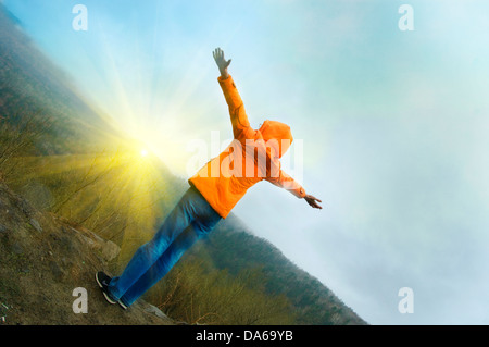 Young woman standing on cliff's edge and looking to a sky with raised hands Stock Photo