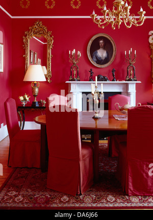 Red loose covers on chairs at oval antique table in bright red dining room with brass chandelier and antique gilt mirror Stock Photo