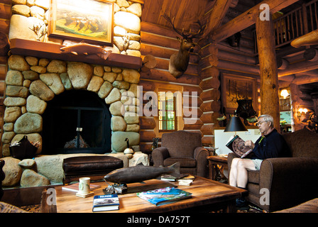 Asian man reading log construction wood plank walls stone fireplace great room Gold River Lodge BC Stock Photo