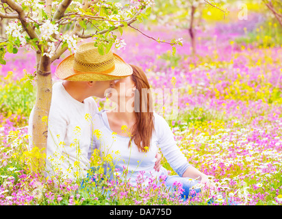Cute young family sitting down on pink floral meadow in the garden and kissing, woman and man enjoying each other outdoors Stock Photo