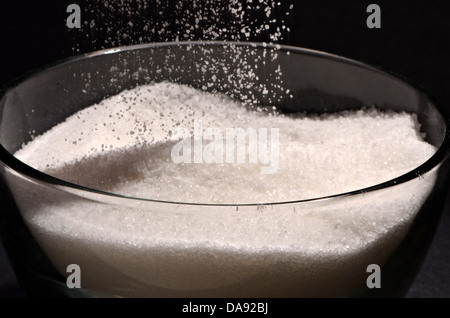 Sugar is falling into a glass sugar bowl on a black background. Stock Photo