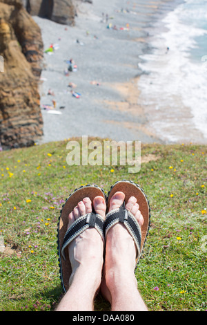Men's feet wearing a pair of flip flop sandals on a cliff top above Sandymouth Bay, Devon, England on a sunny summer day. Stock Photo