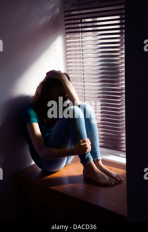 A teenage girl, hand on her head, sitting by a window with light pouring in. Stock Photo