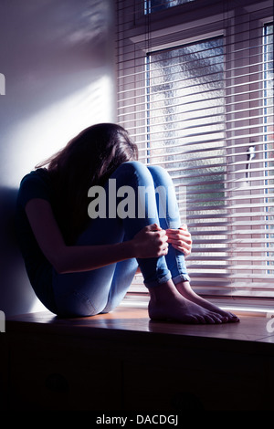 Teenage girl, head bowed, sitting by a window with light pouring in. Stock Photo