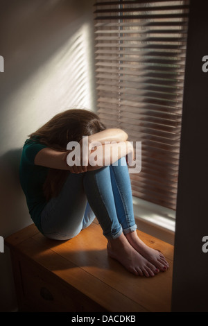 A teenage girl, head down, sitting by a window with light pouring in. Stock Photo