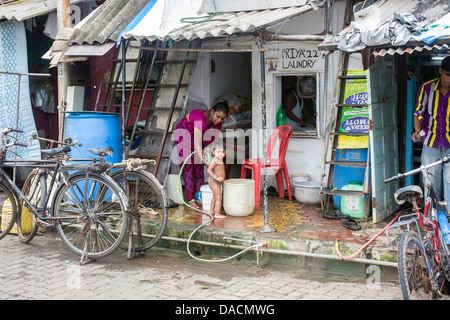 Slum dwellings on Carter Road, Mumbai, small laundry business and home where woman is washing her young child from a bucket Stock Photo