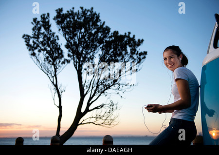 Smiling young woman leaning on camper van at dusk Stock Photo