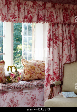 Pink floral curtains and pelmet on open window above window seat with floral and checked cushions in cottage living room Stock Photo