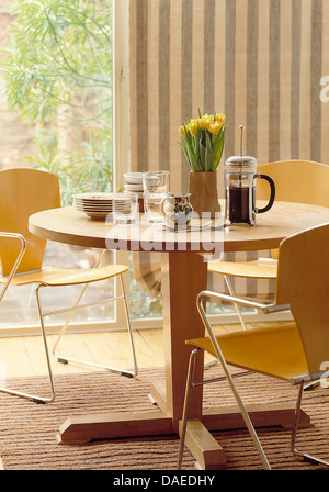 Cafetiere on pale wood circular table with plywood and steel chairs in modern dining room with striped blind on glass doors Stock Photo