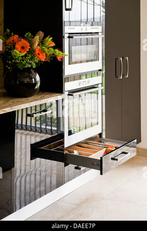 Open cutlery drawer below integral oven in modern kitchen with vase of orange gerberas on worktop Stock Photo