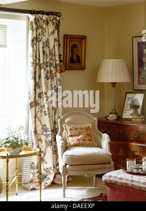 Upholstered French style armchair in front of window with floral curtains in country sitting room Stock Photo