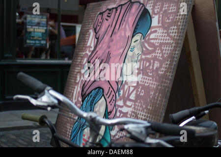 A large pink and blue figurative street-art canvas is displayed behind parked bicycles during a Temple Bar Night Market summer evening event at Crampton Court, Temple Bar, Dublin 2, Ireland, in July 2013. The stylised portrait, shown after or between stages of live creation, stands on a temporary outdoor display while bicycle handlebars cross the foreground. The market brought original canvases, live painting and handmade crafts into the public space as part of Temple Bar’s community arts activity. Stock Photo