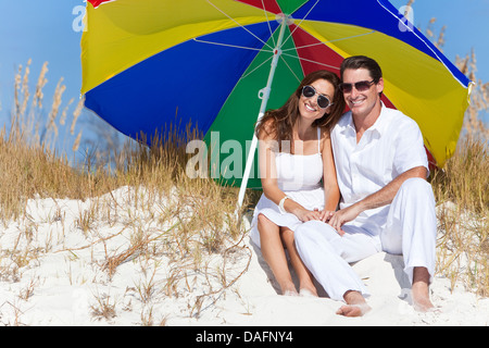 Man and woman romantic couple wearing sunglasses under a multi colored sun umbrella or parasol on a beach Stock Photo