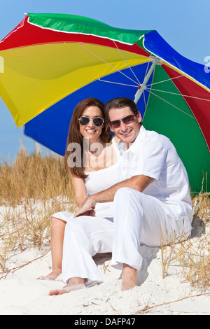 Man and woman romantic couple wearing sunglasses under a multi colored sun umbrella or parasol on a beach Stock Photo