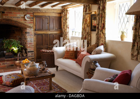 White sofas piled with cushions and rustic wooden coffee table in cottage sitting room with exposed brick fireplace Stock Photo