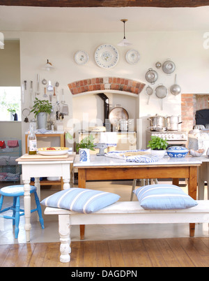 Blue striped cushion on white bench at simple wooden table in country kitchen with display of plates and pans on wall above Aga