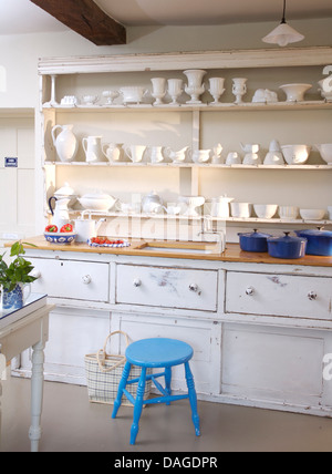 White china displayed on shelves above sink in white built in dresser in cottage kitchen with bright blue stool Stock Photo