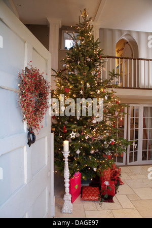 Wreath on open front door with view of tall Christmas tree and gift wrapped presents in double height country hall Stock Photo