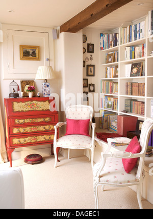 Two period style chairs with red silk cushions in front of fitted bookcase in country living room with painted antique bureau Stock Photo