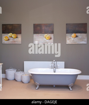 Freestanding roll top bathtub in center of gray traditional bathroom with three white baskets below three paintings of peaches Stock Photo