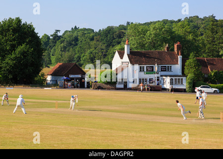 Local teams playing a cricket match on village green with scorched grass in front of Barley Mow pub on a summer evening. Tilford Surrey England UK Stock Photo