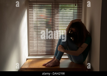 A teenage girl, hand on her head, sitting by a window with light pouring in. Model and property released. Stock Photo