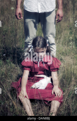 a sad girl sitting on a field, a man is standing behind her Stock Photo