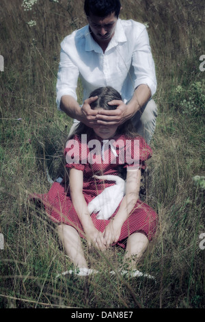 a girl is sitting in the grass, a man is holding his hands in front of her eyes Stock Photo
