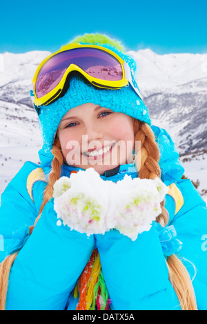 Happy 10 years old girl showing pile of snow wearing beautiful fancy winter clothes and ski mask with mountains on background Stock Photo