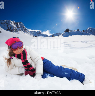 Cute 10 years old girl in ski mask and bright snow clothes laying in snow with mountains on background Stock Photo