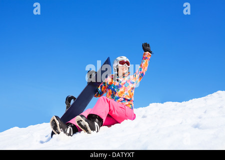 Happy woman in pink sit in snow holding snowboard waiving hands, with mountains on blue sky background Stock Photo