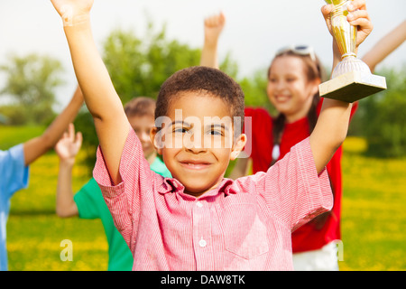 Close shoot of black happy smiling little boy holding prize cup with other kids cheering on the back Stock Photo