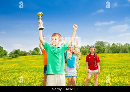 Portrait of happy little boy holding prize in lifted hands cup with his team cheering on background Stock Photo