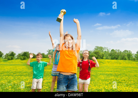 Portrait of happy little girl holding prize in lifted hands cup with her team cheering on background Stock Photo